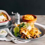 plate piled with enchiladas thermomix pepper grinder old style in the background, along with baking dish of more enchiladas