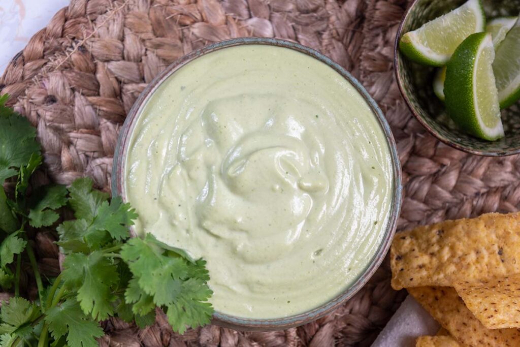 lime coriander cashew sauce in a bowl, on brown placemat. Fresh coriander and lime and corn chips