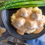 grey tabletop, with black bowl of mashed potato, vegetarian meatballs and Swedish cream sauce gravy, a few pieces of asparagus at back of plate. Small pouring jug bottom left with cream sauce in. Knife and fork and blue serviette front middle and right.