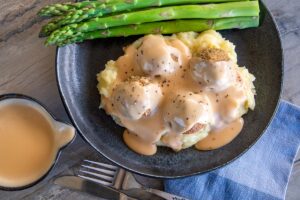 grey tabletop, with black bowl of mashed potato, vegetarian meatballs and Swedish cream sauce gravy, a few pieces of asparagus at back of plate. Small pouring jug bottom left with cream sauce in. Knife and fork and blue serviette front middle and right.