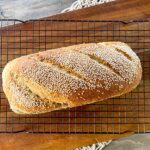 whole loaf super seeded wholemeal multigrain thermomix bread Birdseye view. Loaf has sesame seeds on top and is sitting on top of a black mesh cooling rack on top of a brown wooded bread board on grey countertop