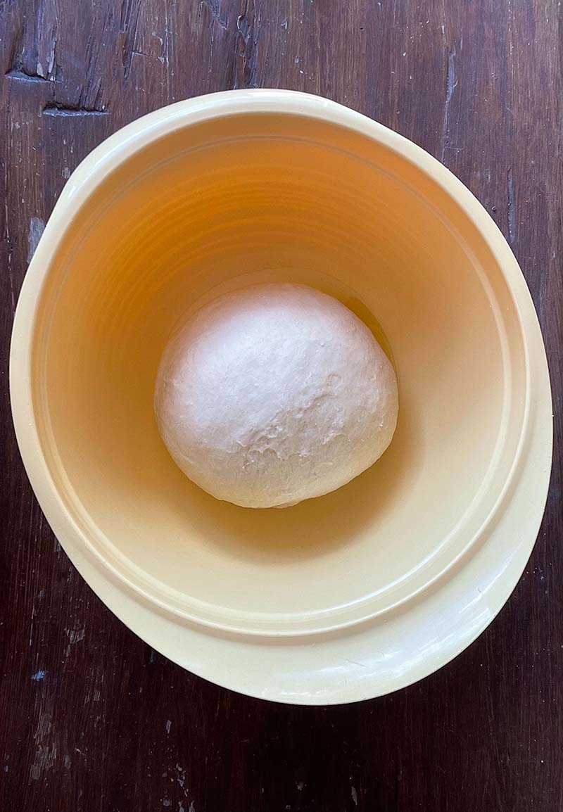 birdseye view of yellow bowl with ball of pizza dough (before rising) in it, sitting on dark brown wooden surface