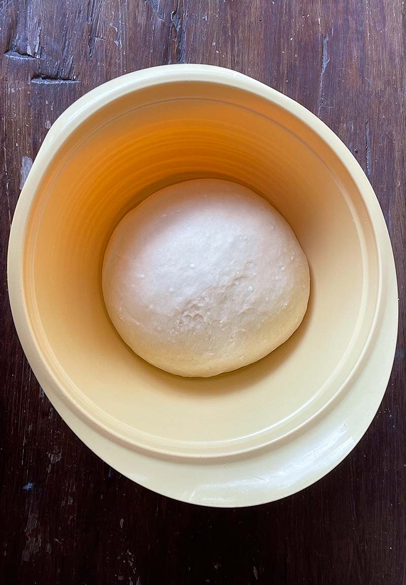 birdseye view of yellow bowl with ball of pizza dough (after rising) in it, sitting on dark brown wooden surface