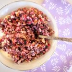 birdseye view shot of a white bowl filled with yoghurt and chopped fruit and nuts. Silver spoon in bowl, bowl sitting on purple serviette with flower pattern