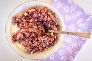 birdseye view shot of a white bowl filled with yoghurt and chopped fruit and nuts. Silver spoon in bowl, bowl sitting on purple serviette with flower pattern