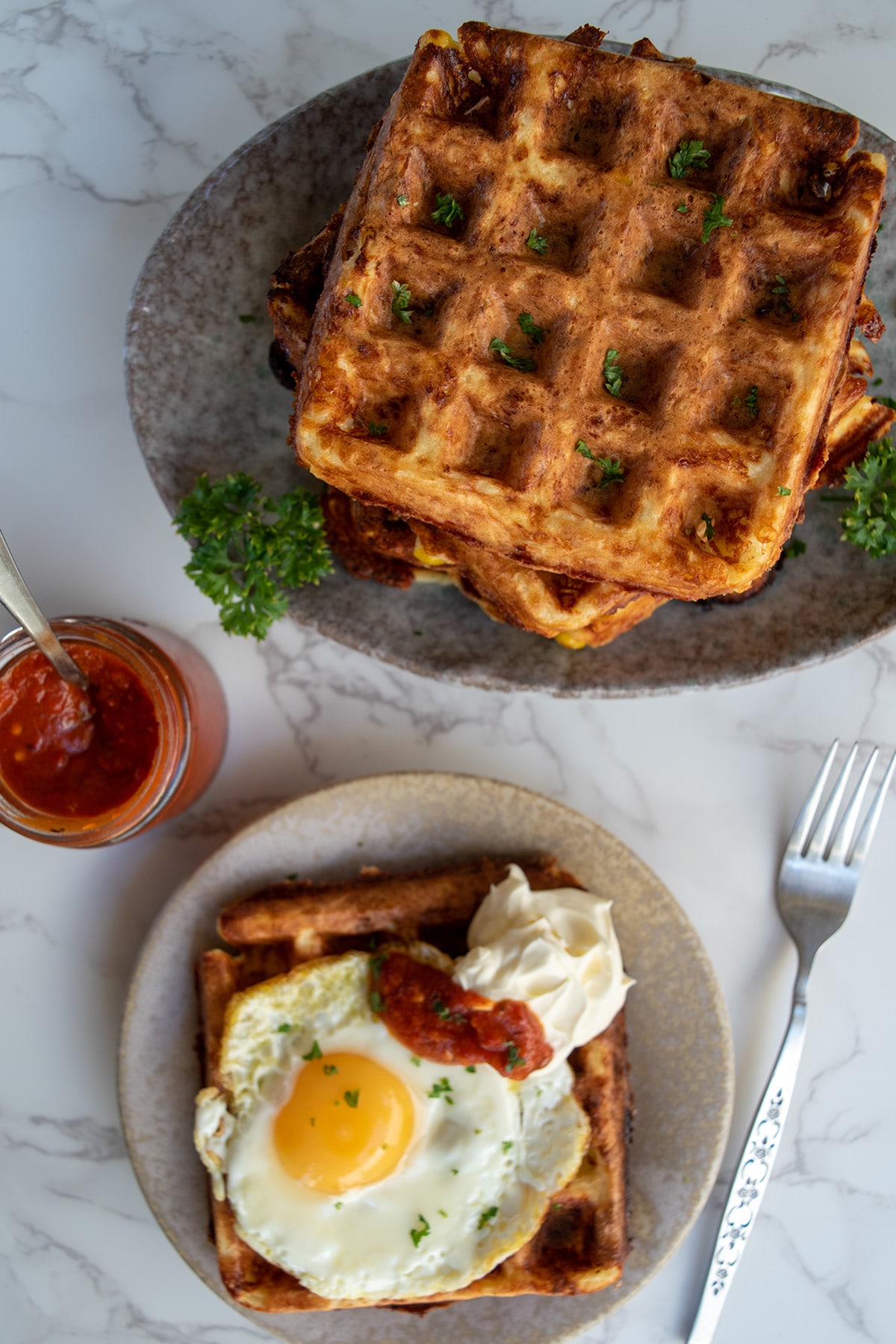 savoury waffles birdseye view two plates, one plated up with a fried egg, sour cream and chilli sauce on top. Jar of chilli sauce and fork next to plate