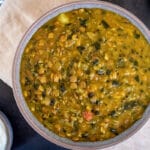 Birdseye view of chunky lentil and spinach soup in grey bowl on cream napkin on black tabletop. Spoon and small bowl of yoghurt with coriander to the left and little dish of coriander to the right