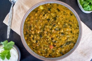 Birdseye view of chunky lentil and spinach soup in grey bowl on cream napkin on black tabletop. Spoon and small bowl of yoghurt with coriander to the left and little dish of coriander to the right