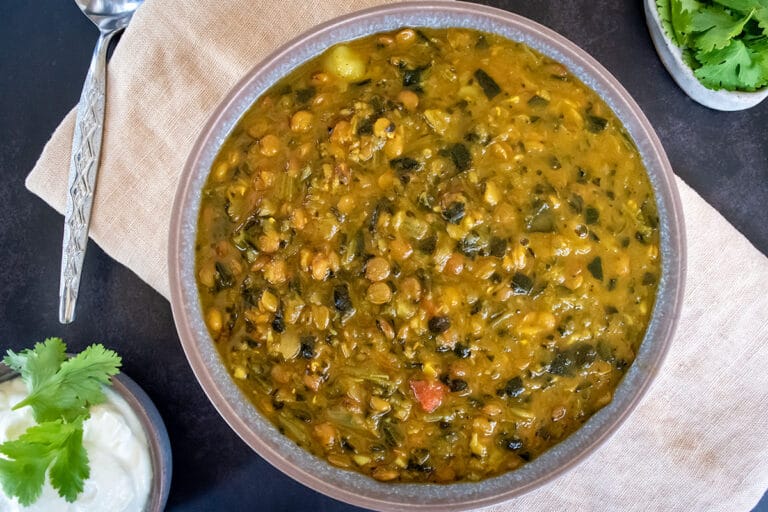 Birdseye view of chunky lentil and spinach soup in grey bowl on cream napkin on black tabletop. Spoon and small bowl of yoghurt with coriander to the left and little dish of coriander to the right
