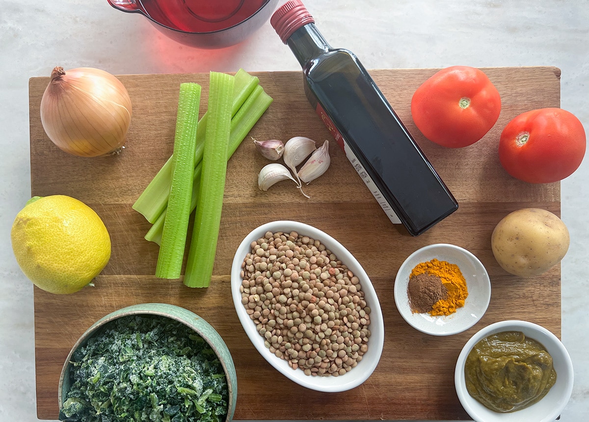 brown chopping board on white bench top, on top of which is onion, celery, garlic, olive oil bottle, tomatoes, potato, stock paste, spices, lentils, spinach, lemon - ingredients are for lentil spinach soup in thermomix