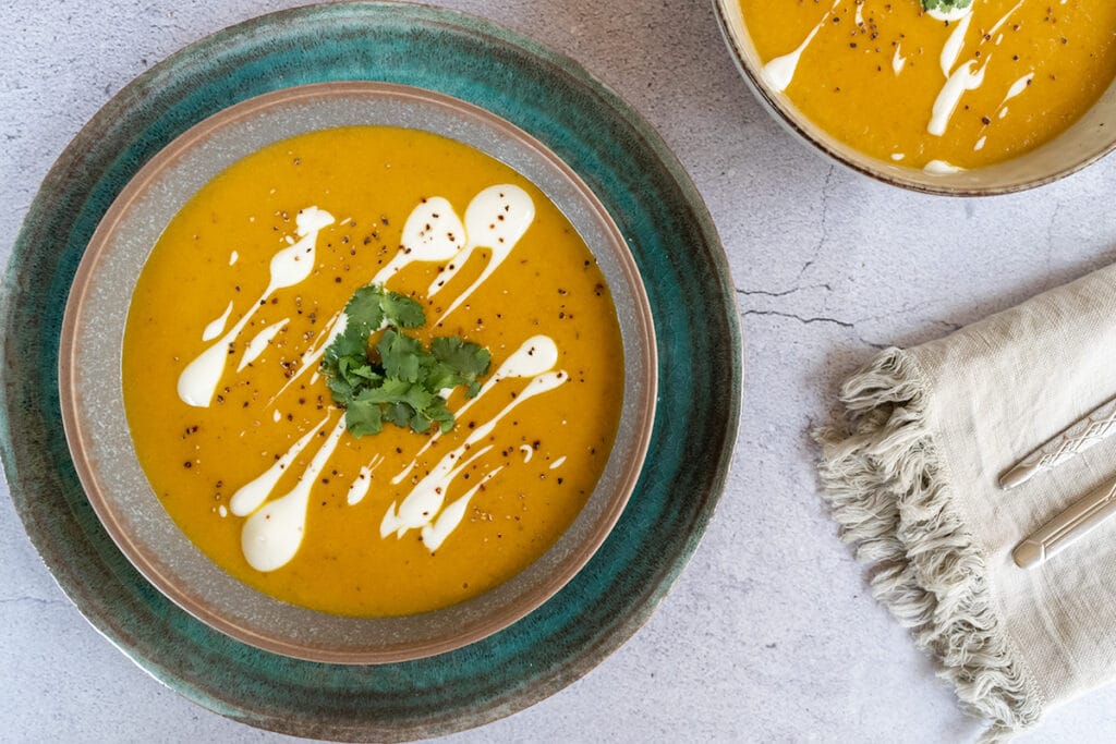 bowl of orange coloured sweet potato soup, topped with yoghurt swirls, coriander and pepper. Grey table top