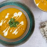 bowl of orange coloured sweet potato soup, topped with yoghurt swirls, coriander and pepper. Grey table top