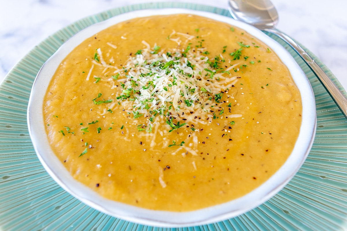 potato and leek soup in white bowl on green plate with spoon at an angle on the plate. Soup is topped with Parmesan, pepper and chopped parsley