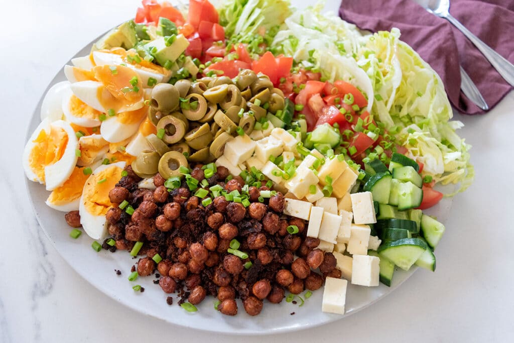 cobb salad on large white plate on white table, maroon napkin and serving utensils in background