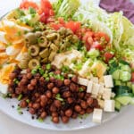 cobb salad on large white plate on white table, maroon napkin and serving utensils in background