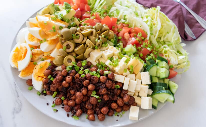 cobb salad on large white plate on white table, maroon napkin and serving utensils in background