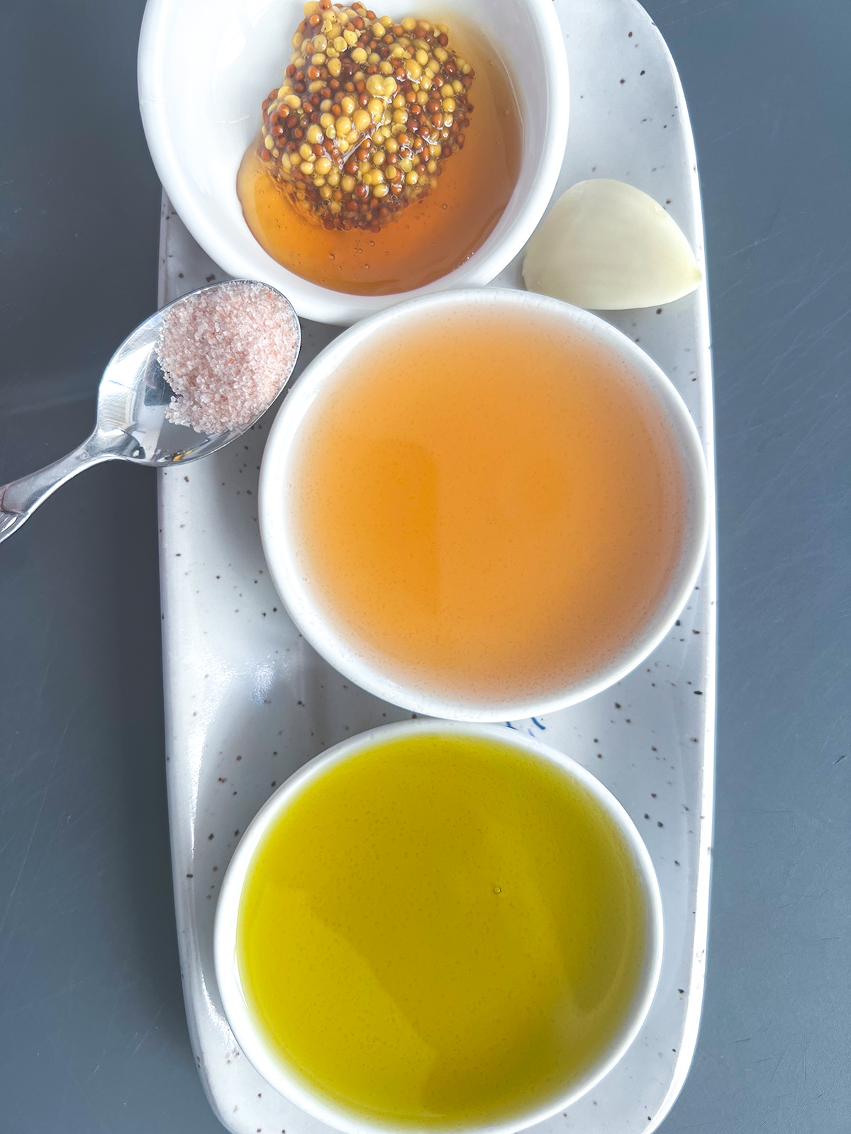 three white ramekins containing oil, vinegar, honey and mustard, and a clove of garlic for couscous salad dressing