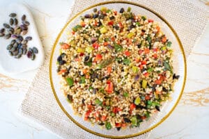 festive couscous salad made in thermomix, in glass bowl with gold rim. Sitting on cream and gold placemat and tabletop. Small white rectangular dish to the top left with toasted pepitas and pistachios in