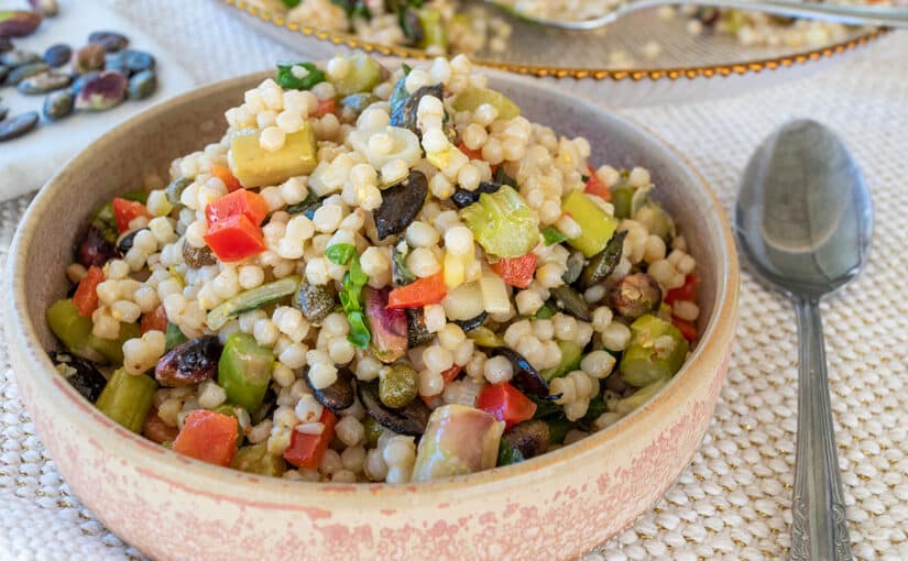 close up of festive couscous salad thermomix in cream and terracotta tapas bowl with spoon to the right. Serving dish containing salad to the back and pistachios between the two dishes