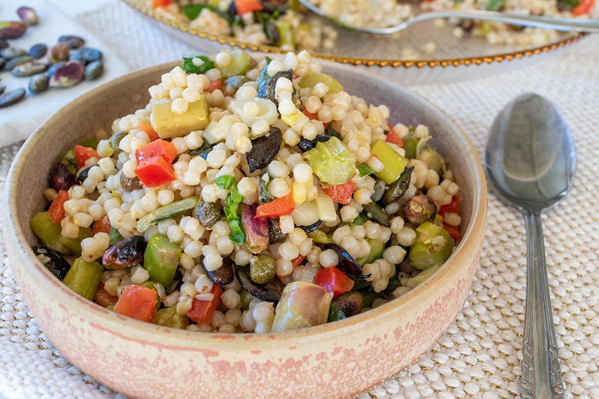 close up of festive couscous salad thermomix in cream and terracotta tapas bowl with spoon to the right. Serving dish containing salad to the back and pistachios between the two dishes