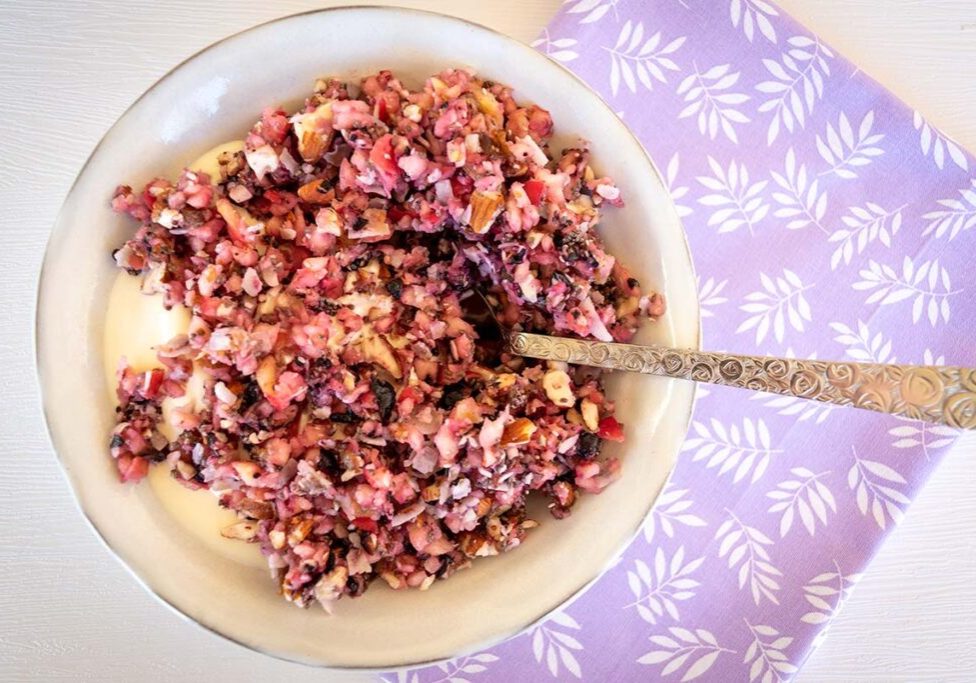 birdseye view shot of a white bowl filled with yoghurt and chopped fruit and nuts. Silver spoon in bowl, bowl sitting on purple serviette with flower pattern