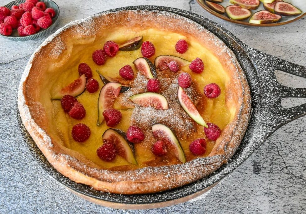 dutch baby pancake in black cast iron pan on rustic background. Fruit inside and outside on plates