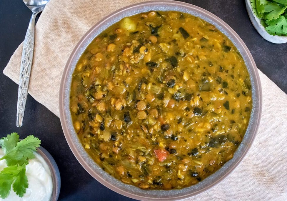 Birdseye view of chunky lentil and spinach soup in grey bowl on cream napkin on black tabletop. Spoon and small bowl of yoghurt with coriander to the left and little dish of coriander to the right