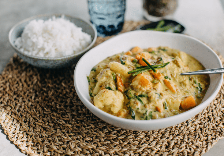 thermomix vegetarian curry in white bowl on straw round placemat with bowl of rice and glass of water in background